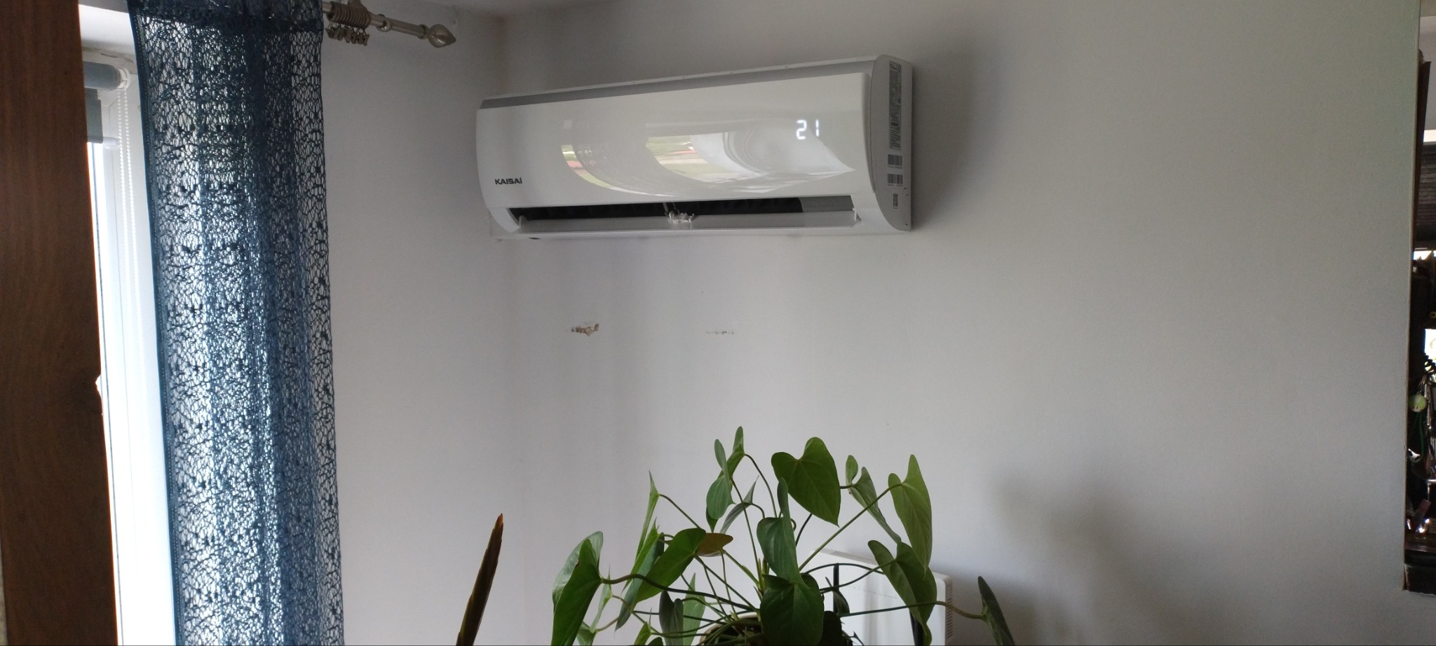 Biały klimatyzator Kaisai zamontowany na ścianie w pomieszczeniu, widoczny wyświetlacz z temperaturą 21 stopni Celsjusza, w tle okno z zasłoną i roślina doniczkowa.