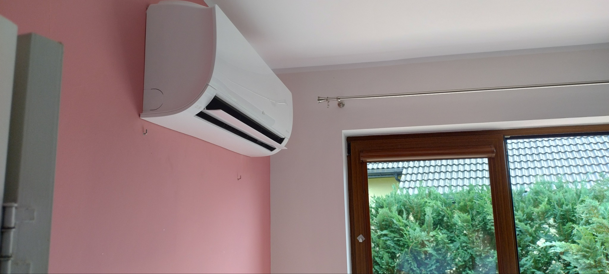 Biała klimatyzacja LG zamontowana na różowej ścianie w pokoju, widok z boku, okno z brązową ramą i zielenią na zewnątrz.