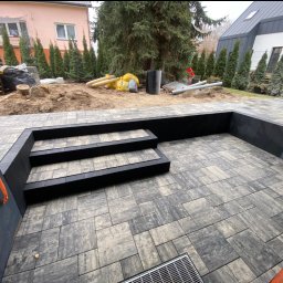 Volodymyr Yarmyshko - Nowoczesny taras z kostki brukowej w odcieniach szarości, z trzema stopniami i kratką odpływową. W tle widoczne przygotowania do dalszych prac ogrodowych.