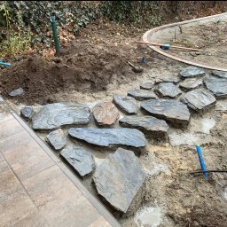 Volodymyr Yarmyshko - Fragment układania ścieżki z nieregularnych płyt kamiennych w ogrodzie. Częściowo ułożona nawierzchnia, widoczne narzędzia i przygotowane podłoże.