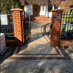 Volodymyr Yarmyshko - Ułożony z kostki brukowej, zakrzywiony chodnik prowadzi do domu z otwartą, czarną bramą. Czerwone, ceglane słupki bramy i obrzeża chodnika. Jesienne liście na pnączach domu.