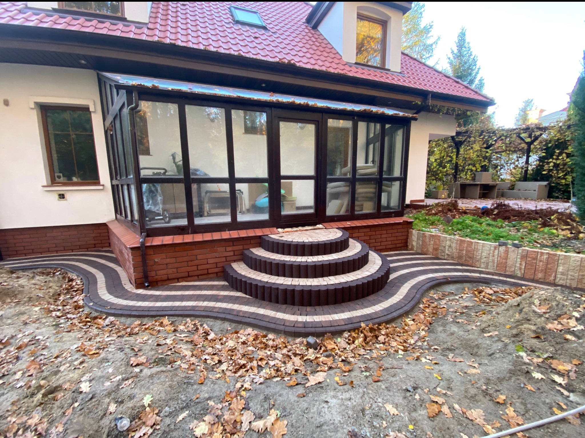 Falista ścieżka z kostki brukowej prowadząca do domu z werandą. Schody z kostki w kształcie półkola. Jesienne liście na ziemi. Widok na ogród i elementy architektury.