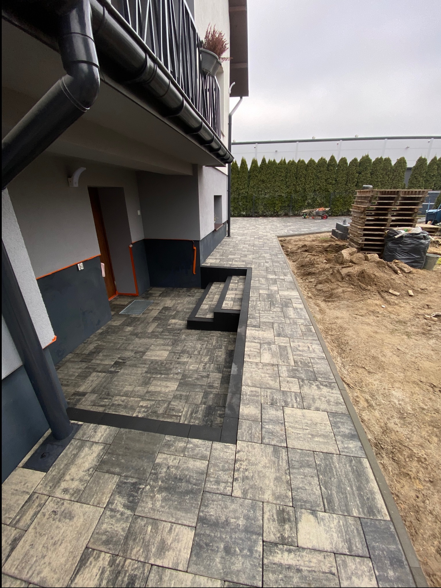 Ułożona kostka brukowa w Warszawie, tworząca ścieżkę i schody przed domem. Widoczne palety z kostką i przygotowany teren pod dalsze prace.