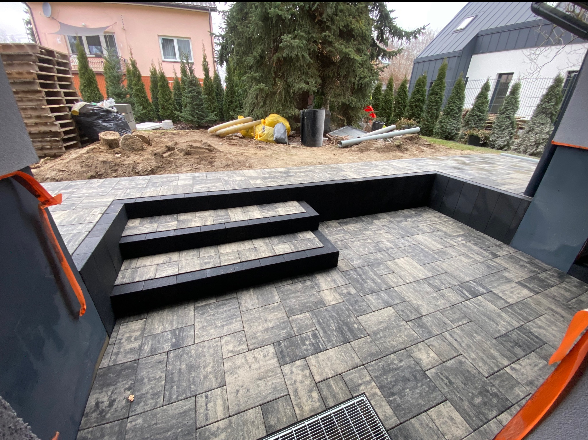 Nowoczesny taras z kostki brukowej w odcieniach szarości, z trzema stopniami i kratką odpływową. W tle widoczne przygotowania do dalszych prac ogrodowych.