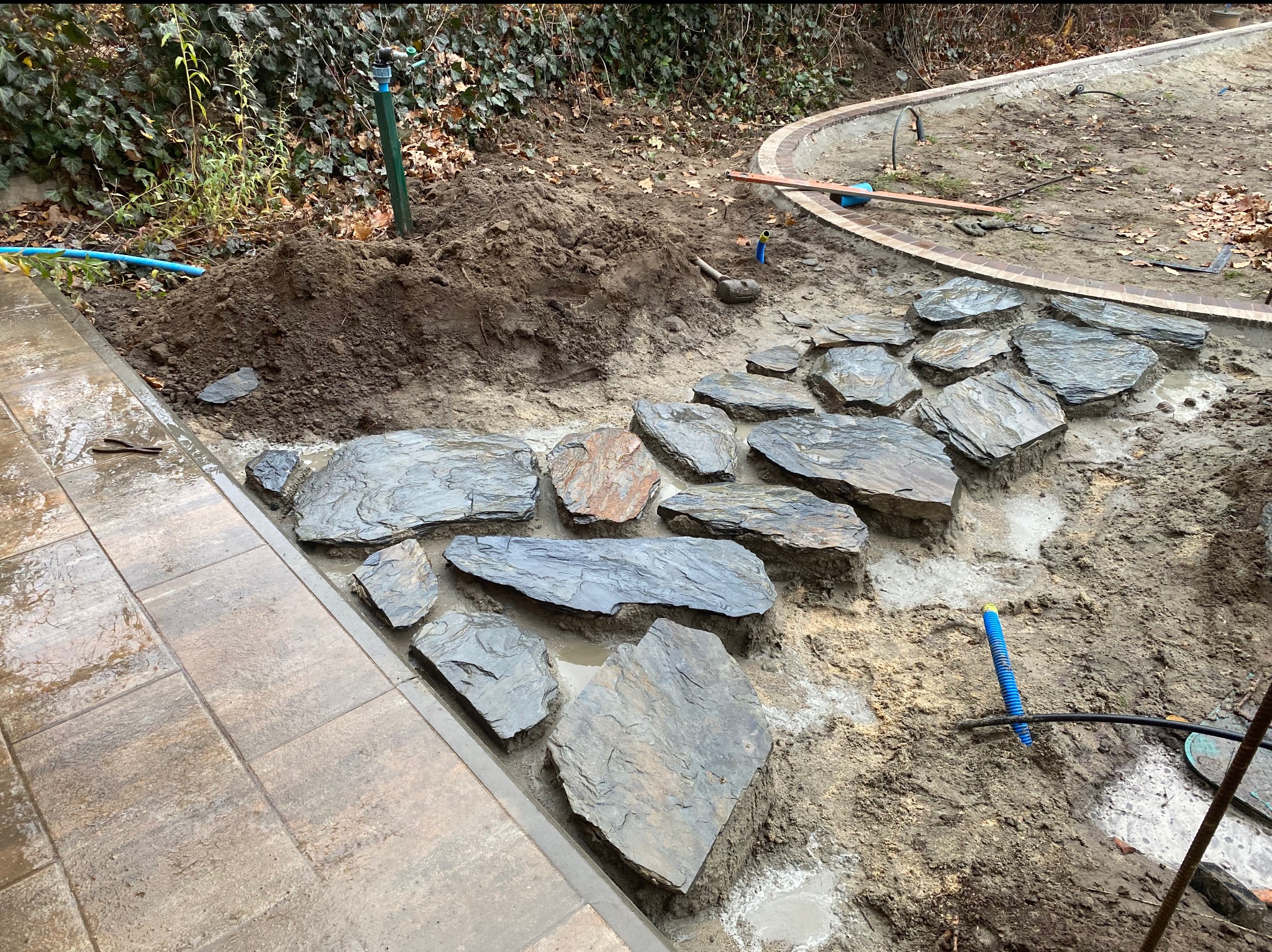 Fragment układania ścieżki z nieregularnych płyt kamiennych w ogrodzie. Częściowo ułożona nawierzchnia, widoczne narzędzia i przygotowane podłoże.