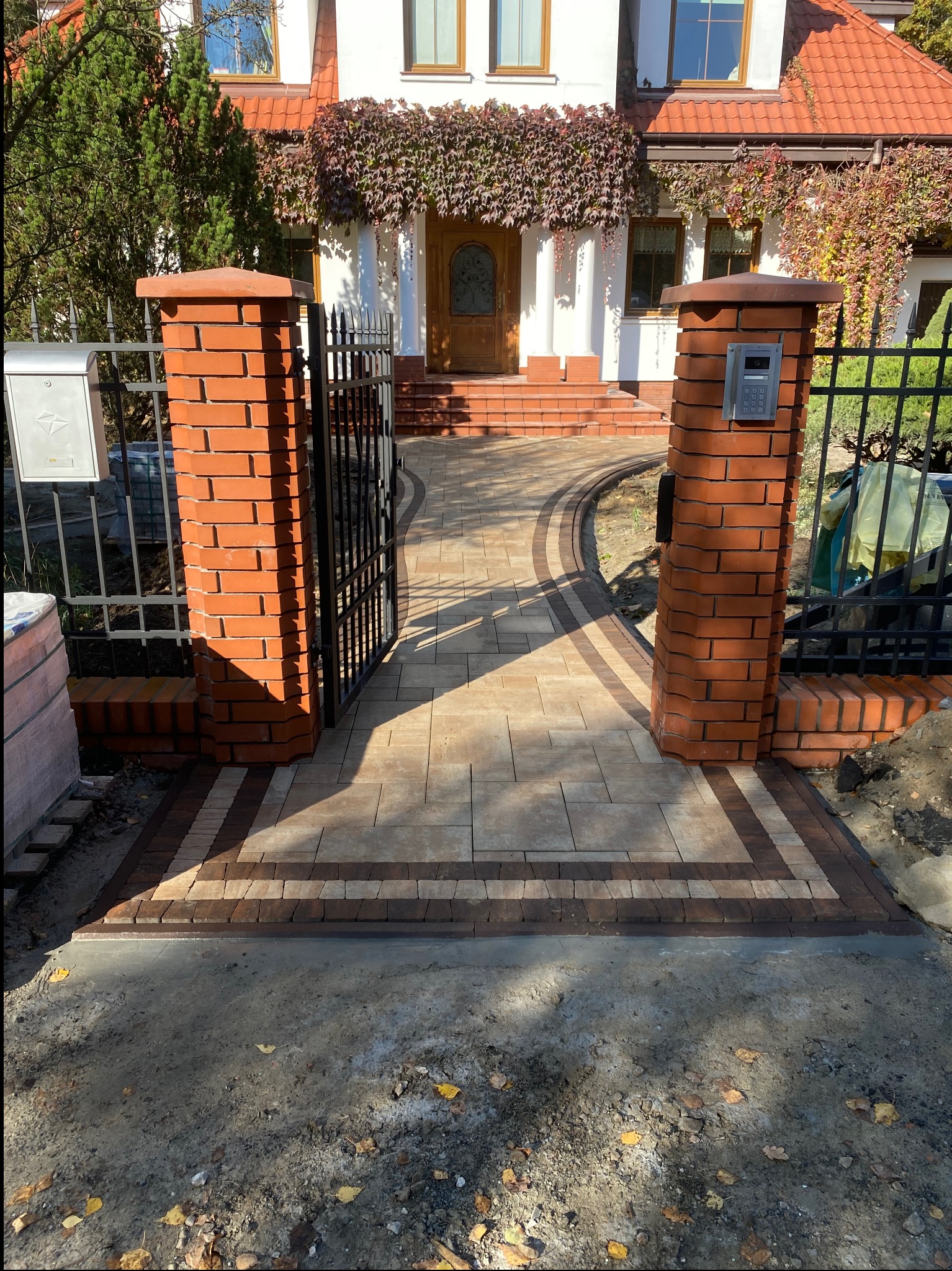 Ułożony z kostki brukowej, zakrzywiony chodnik prowadzi do domu z otwartą, czarną bramą. Czerwone, ceglane słupki bramy i obrzeża chodnika. Jesienne liście na pnączach domu.