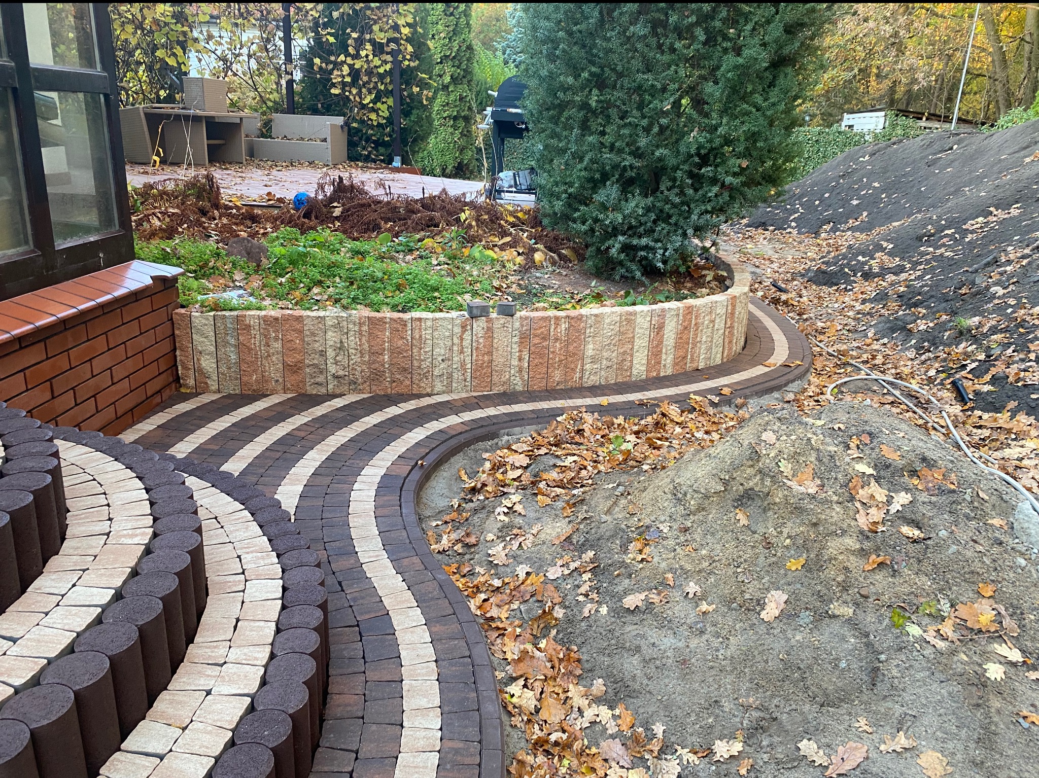 Ogród z ścieżką z kostki w odcieniach brązu i beżu, prowadzącą od schodów do rabaty z kamienną obudową. W tle drzewa i elementy wyposażenia ogrodu.
