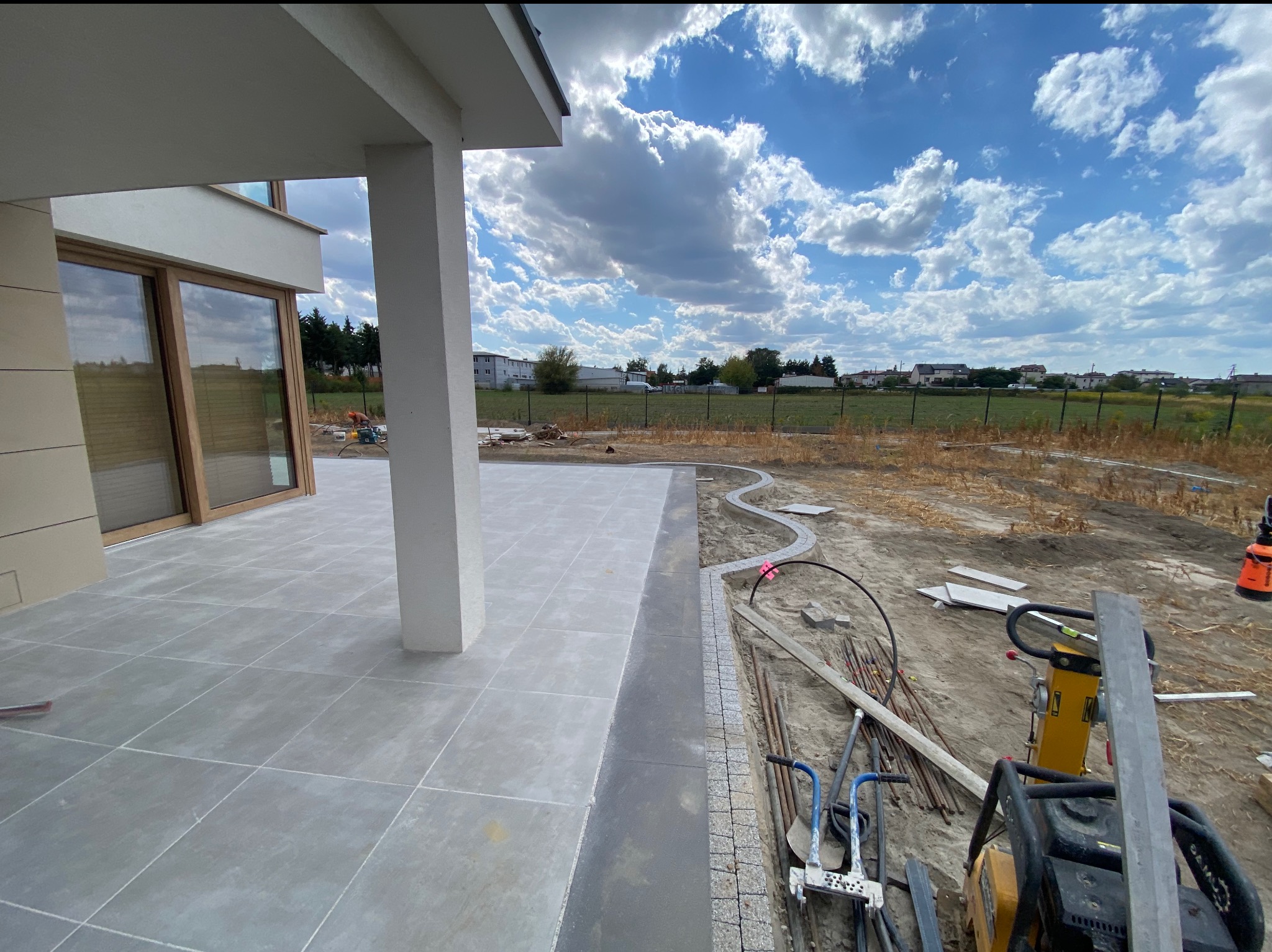 Jasnoszara, ułożona kostka na tarasie domu jednorodzinnego. W tle niebo z chmurami i teren przygotowany pod dalsze prace brukarskie. Widoczne narzędzia i materiały.