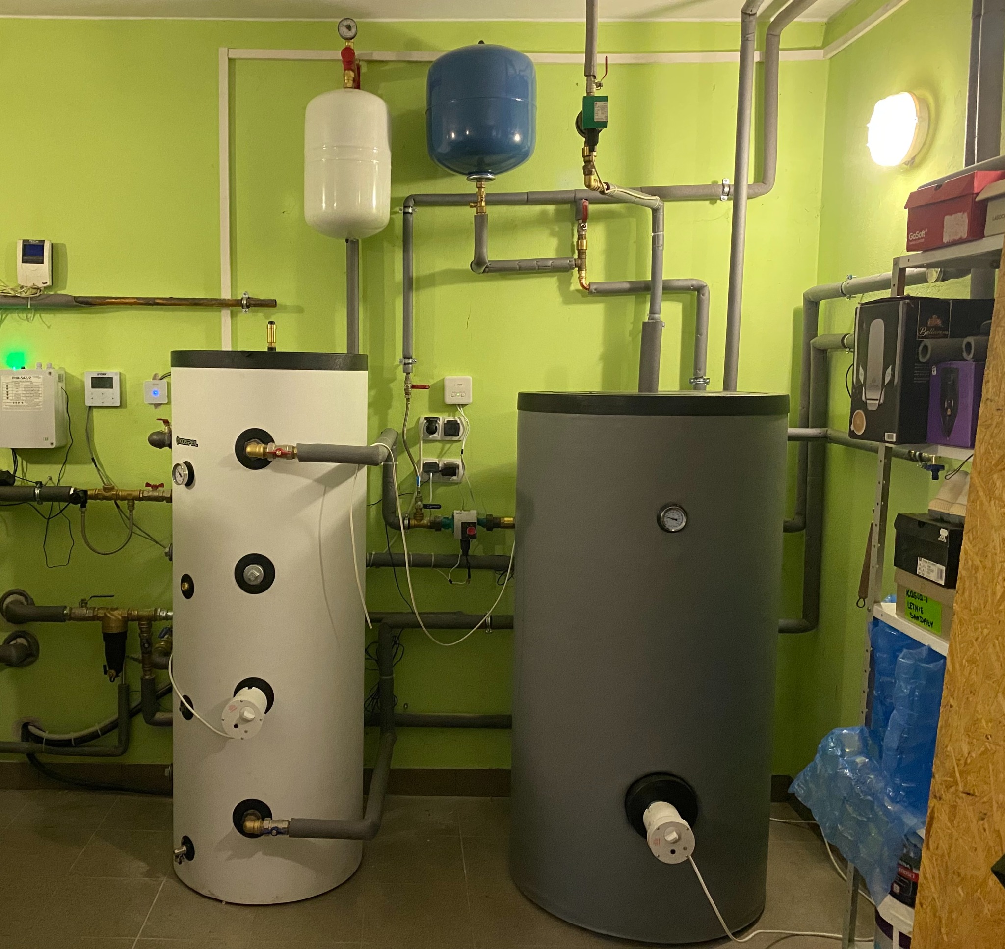 Zestaw instalacji CO w kotłowni z białym buforem Kospel 200l, szarym zasobnikiem CWU Fish 400l i UPS jako systemem przeciwzamrożeniowym na tle zielonej ściany.