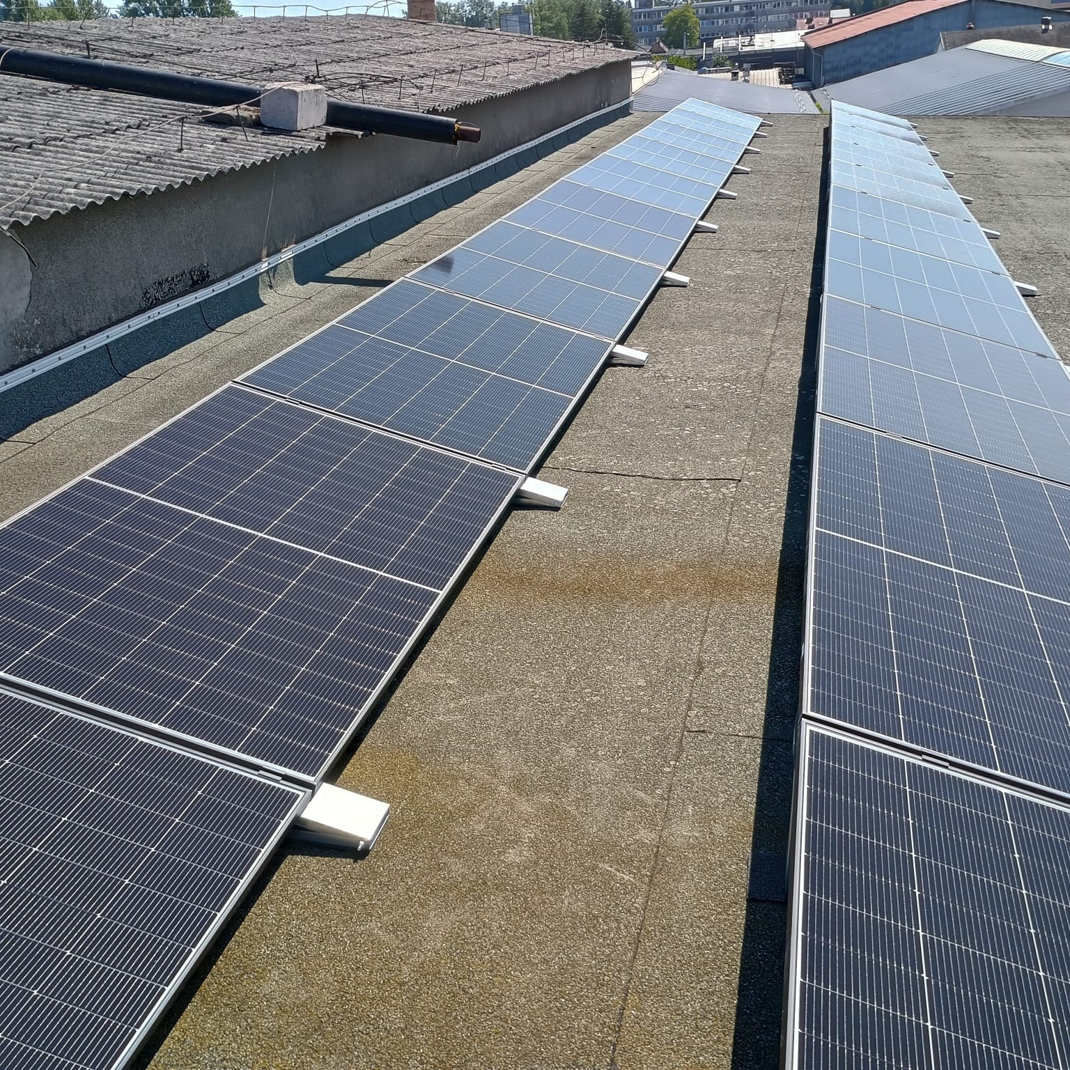 Rząd paneli fotowoltaicznych na dachu budynku, widok z góry. Szara, płaska powierzchnia dachu, panele ustawione w równych odstępach, w tle fragmenty innych budynków.