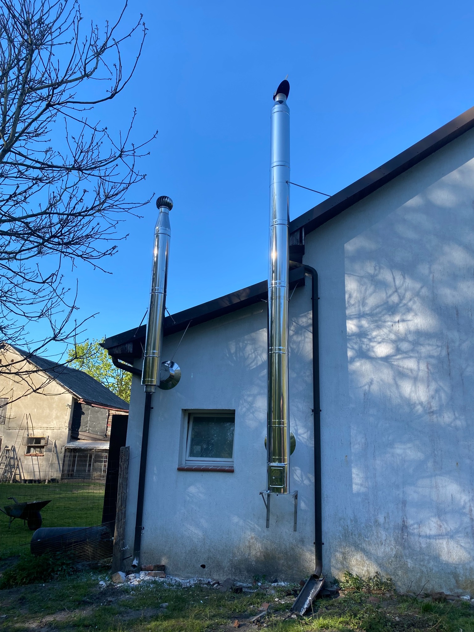 Dwa kominy stalowe, jeden z turbiną wiatrową, zamontowane na ścianie budynku. Widoczne rury spustowe i fragment drzewa. Błękitne niebo w tle.