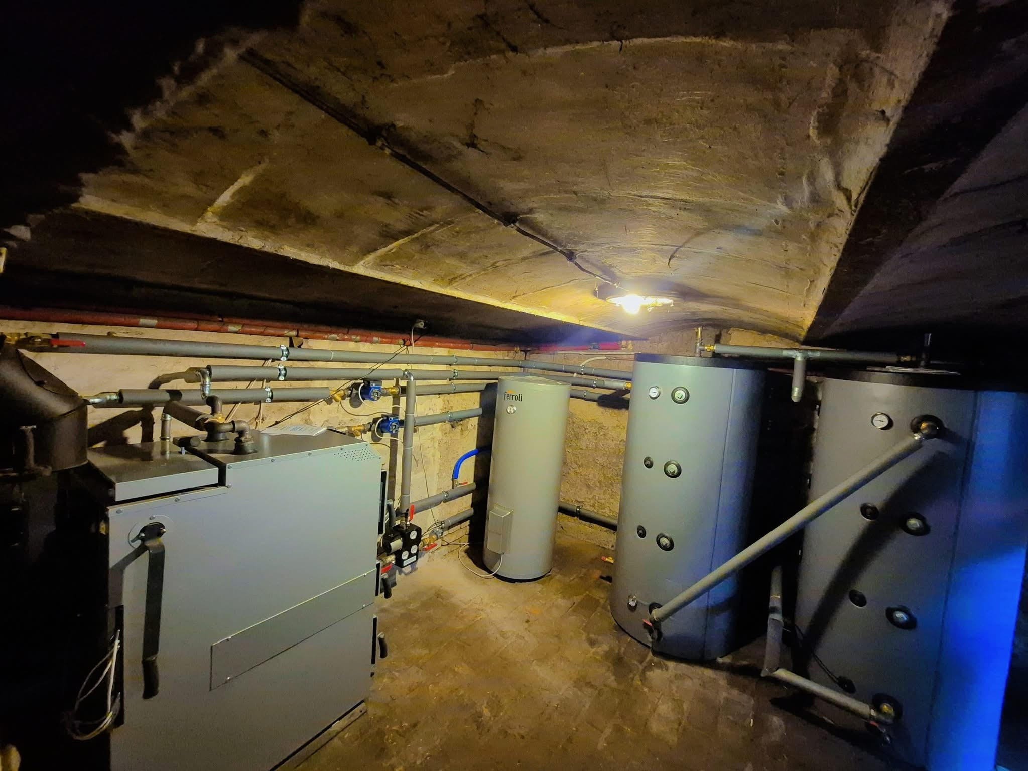 Kotłownia: zbiorniki buforowe (2x500L), CWU 200L z grzałką i Laddomat w piwnicy ze sklepieniem łukowym. Instalacja grzewcza w szarej tonacji, widoczne rury i zawory.