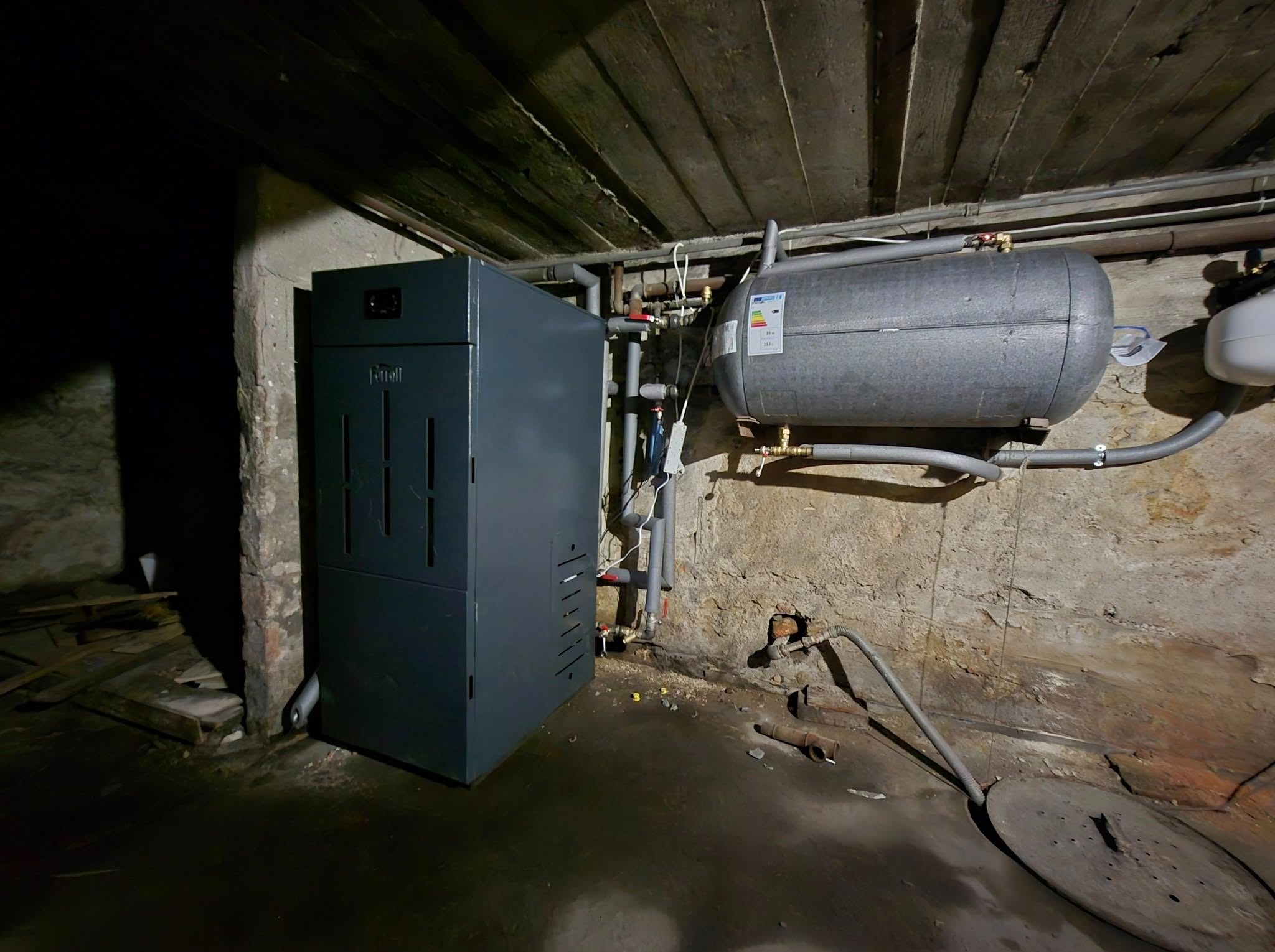 Szara kotłownia z piecem Ferroli Biopellet Pro 12 kW, zasobnikiem Cwu Trinity 120 litrów i sterownikiem Salus. Instalacja w piwnicy z rurami i drewnianym sufitem.