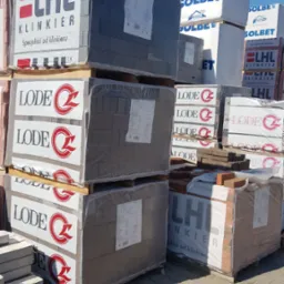 Palety z zapakowanymi cegłami klinkierowymi marek LHL i LODE oraz bloczkami Solbet, składowane na placu budowlanym w słoneczny dzień.