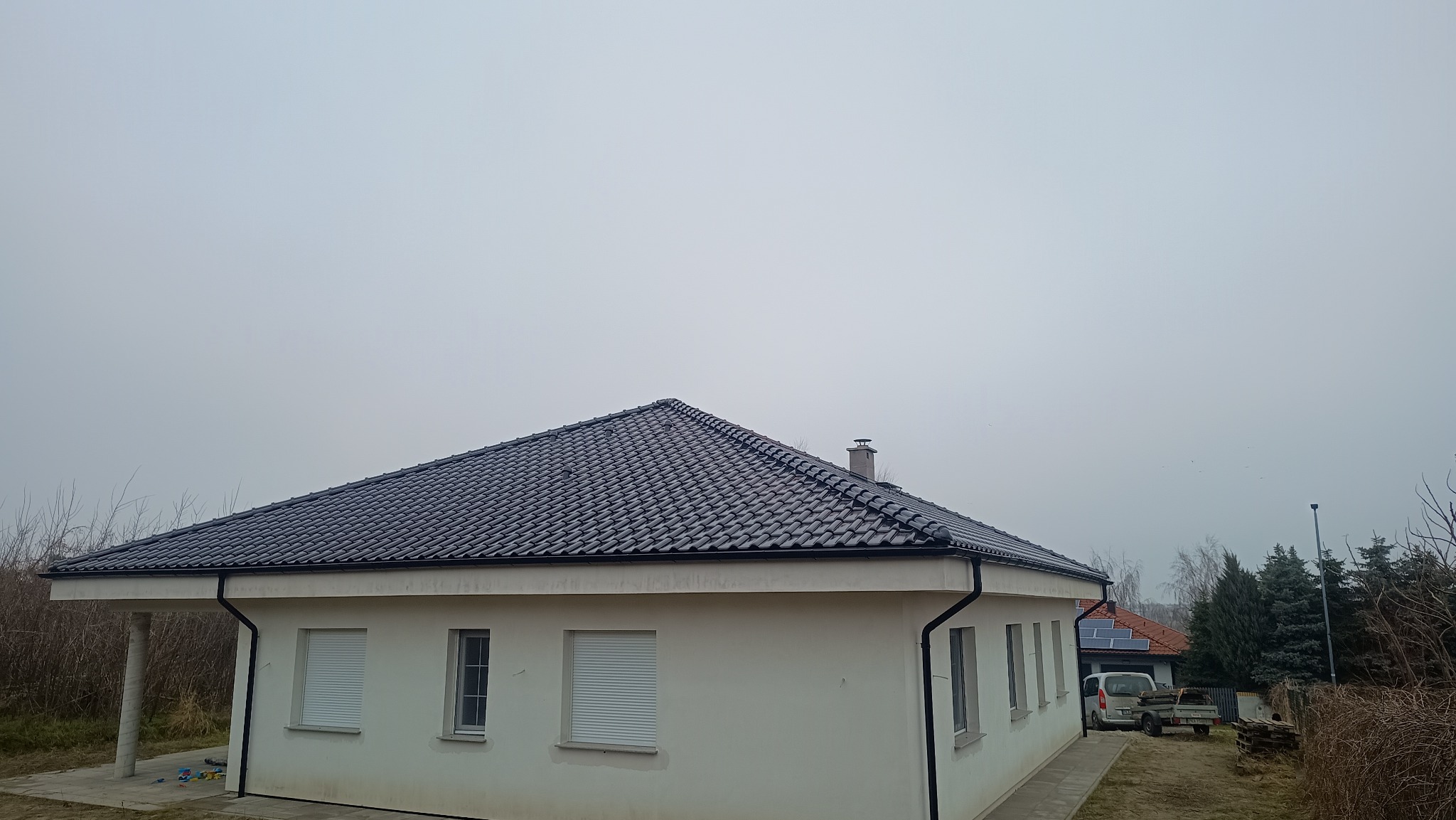 Dom jednorodzinny z ciemną, błyszczącą dachówką ceramiczną i nowymi rynnami. Widoczne okna z roletami i fragment otoczenia w pochmurny dzień.