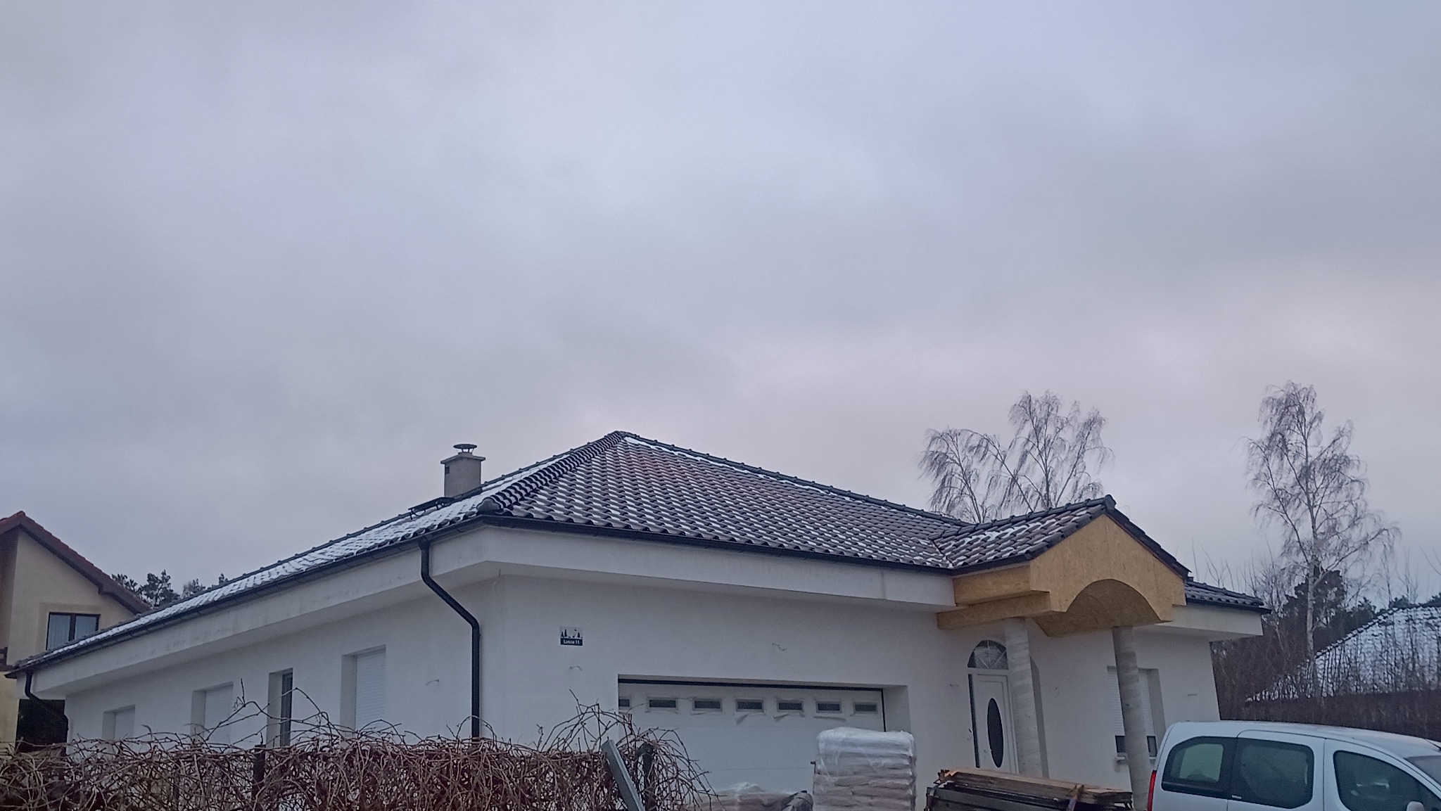 Biały dom z ciemną dachówką w zimowej scenerii. Na dachu leży cienka warstwa śniegu. Widoczne worki z materiałami budowlanymi. Pochmurne niebo w tle.