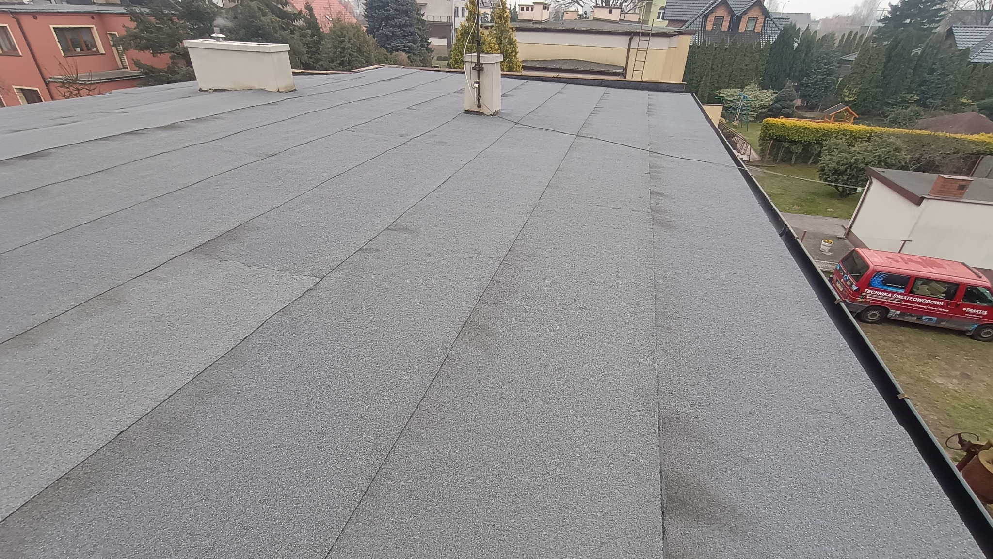 Szary, płaski dach pokryty papą termozgrzewalną z kominem wentylacyjnym. W tle budynki mieszkalne i firmowy samochód dostawczy.