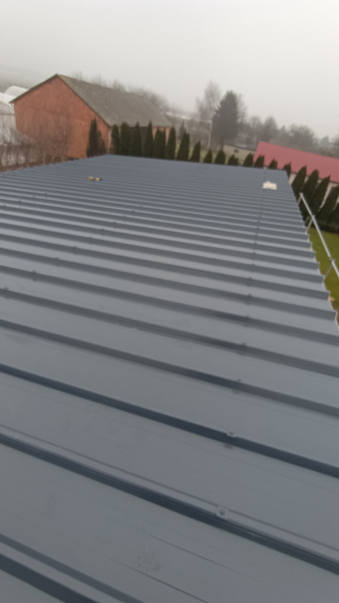 Szary dach z blachy trapezowej, widok z góry na tle zabudowań i zieleni. Widoczne mocowania blachy. Pochmurna pogoda.