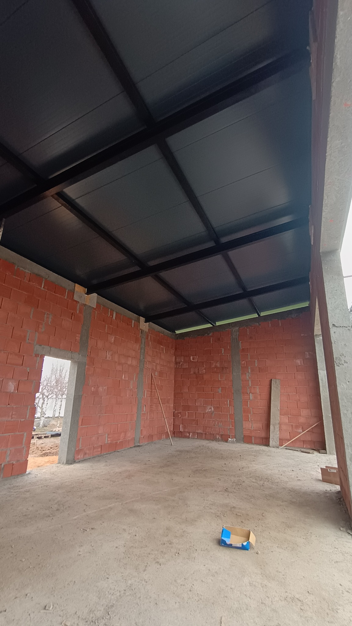 Wnętrze hali w trakcie budowy: surowe ściany z cegły, betonowe słupy i stalowa konstrukcja dachu z szarym pokryciem. Na podłodze leży karton.