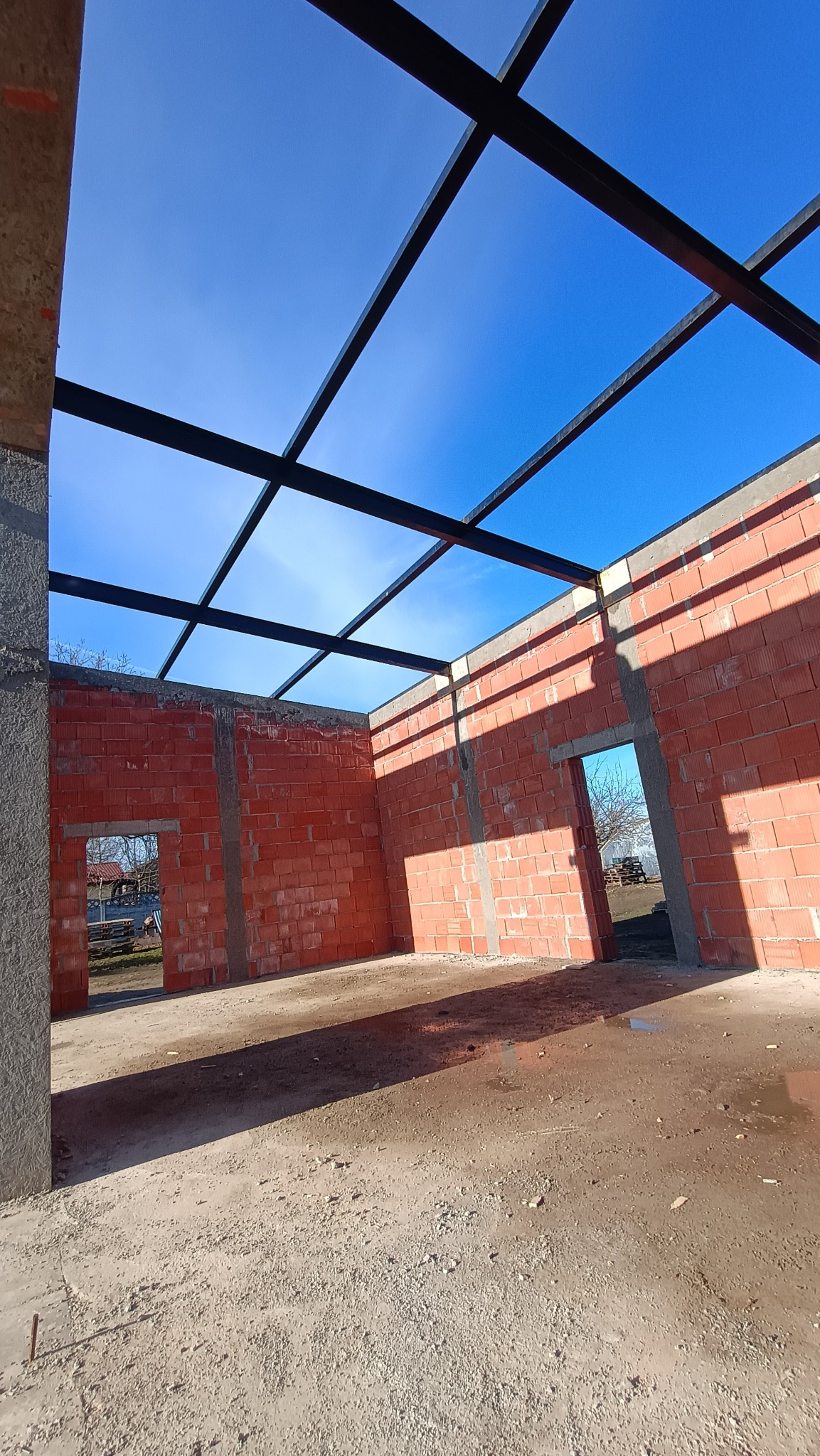 Wnętrze budynku w trakcie budowy z czerwonej cegły, widoczna stalowa konstrukcja dachu na tle błękitnego nieba. Surowy stan, widoczne otwory okienne i drzwiowe.