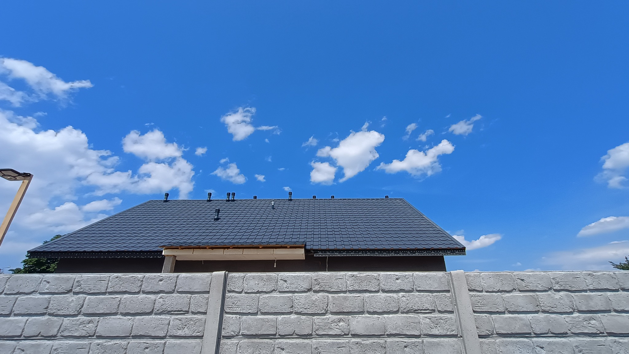 Grafitowy dach z kominkami wentylacyjnymi, widoczny zza betonowego ogrodzenia w słoneczny dzień. Fragment niezakończonej obróbki okapu. Nowoczesne oświetlenie uliczne.