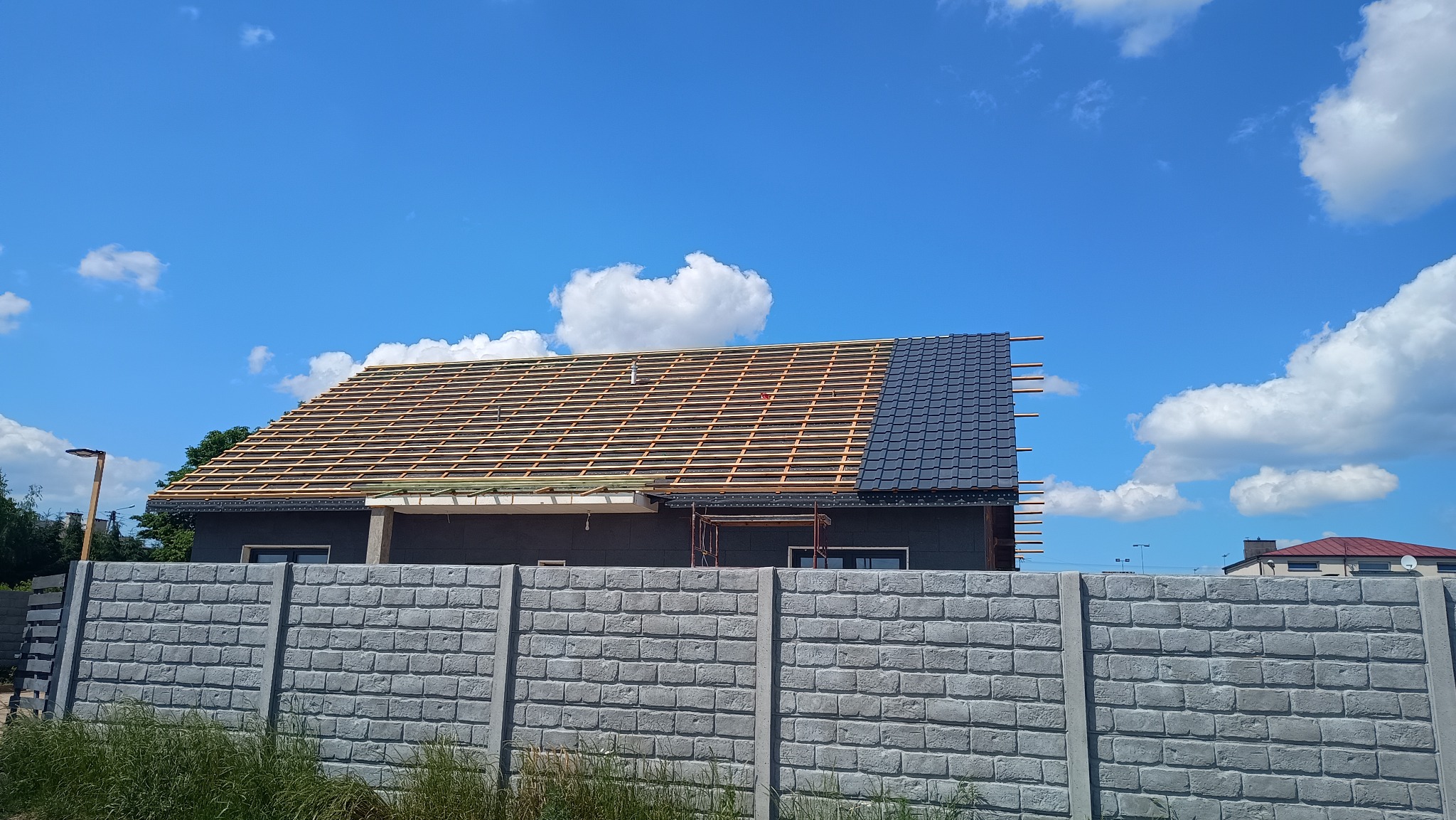 Dom w trakcie krycia dachu: widoczna więźba i fragment pokryty ciemną dachówką. Na pierwszym planie betonowe ogrodzenie, w tle błękitne niebo z chmurami.