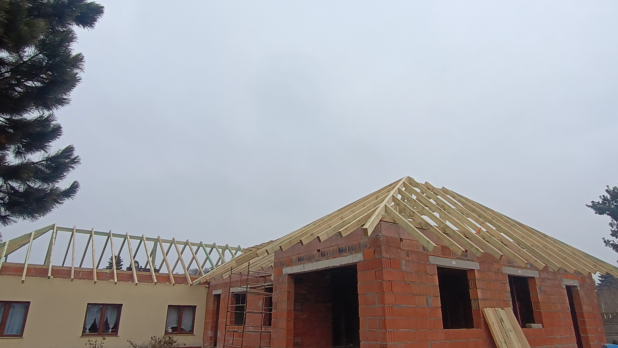 Wznoszenie więźby dachowej na murowanym domu jednorodzinnym w trakcie budowy. Widoczne drewniane elementy konstrukcji na tle pochmurnego nieba.