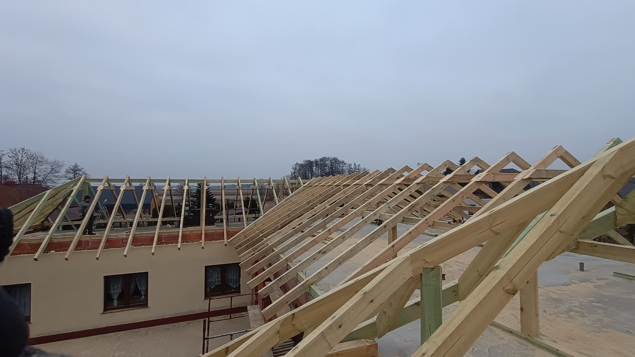 Wznoszenie drewnianej więźby dachowej na murowanym budynku w pochmurny dzień. Widoczne elementy konstrukcyjne i fragmenty otoczenia.