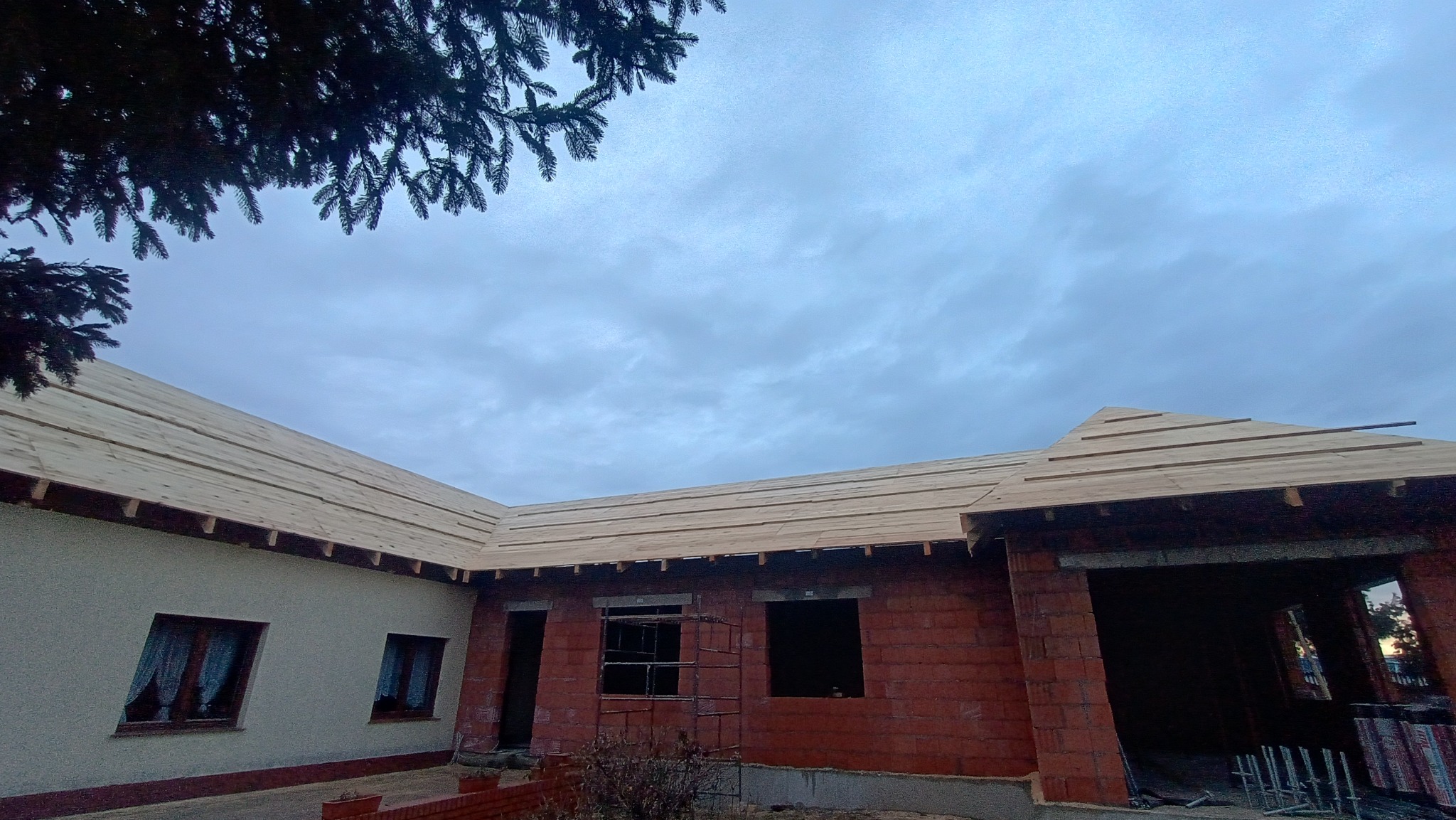 Budowa dachu domu jednorodzinnego z widoczną więźbą i deskowaniem, ceglany mur w tle, pochmurne niebo. Etap prac dekarskich.