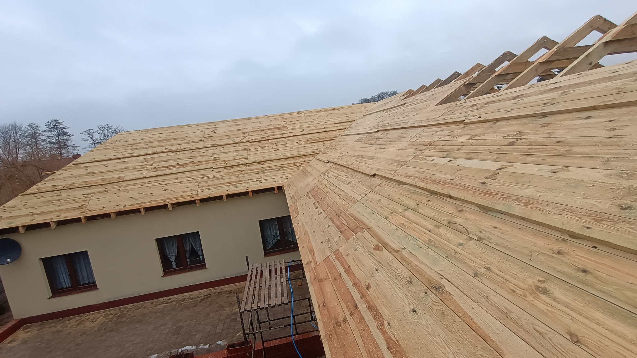 Drewniana konstrukcja dachu w trakcie budowy, widok z góry na deski i krokwie, dom w tle, pochmurne niebo. Widoczny fragment rusztowania.