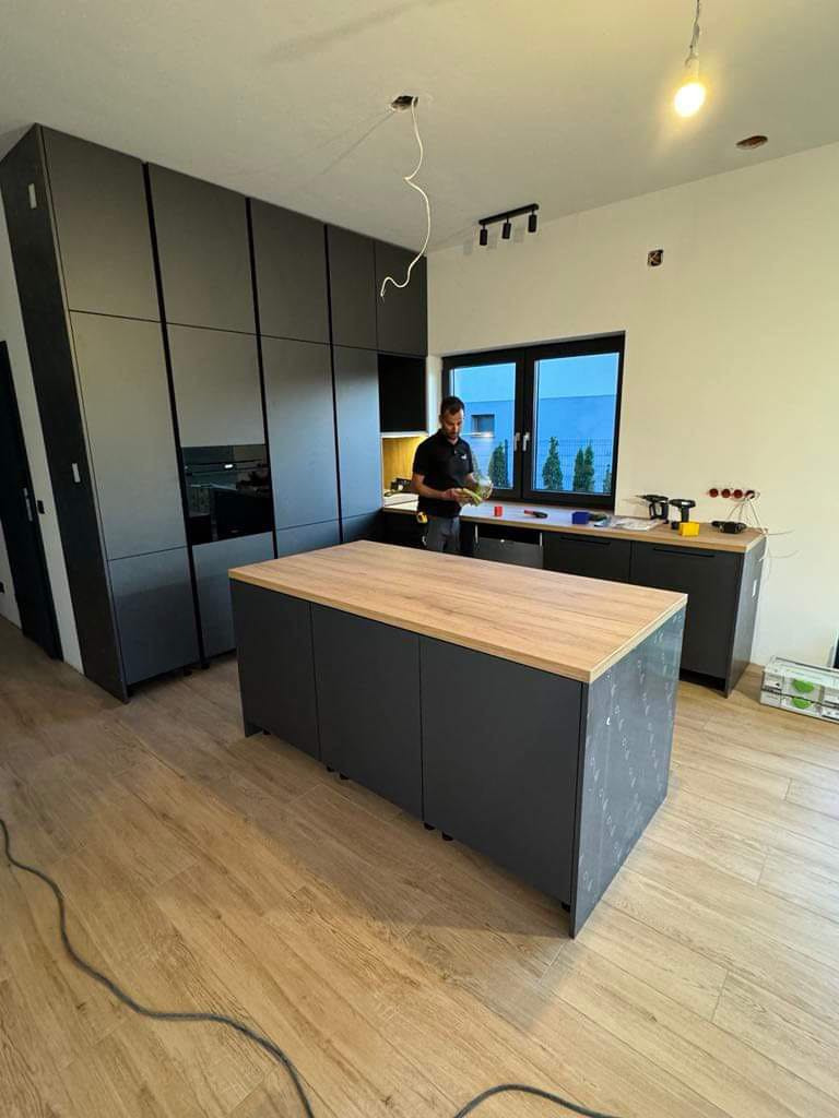 Montaż nowoczesnej, szarej kuchni z drewnianym blatem w Szczecinie, widoczny mężczyzna podczas pracy, na podłodze panele.