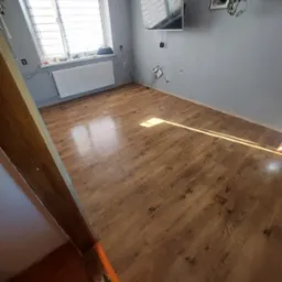 Ułożone panele podłogowe w pokoju z szarą ścianą, białym grzejnikiem i telewizorem na ścianie. Widoczny fragment futryny drzwi.