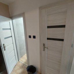 MLB Services - Wnętrze mieszkania w trakcie prac wykończeniowych: widoczne dwie białe, nowoczesne drzwi z poziomymi wstawkami, czarne włączniki światła na ścianie, wiadro na podłodze z terakoty imitującej kamień,...