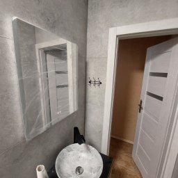 MLB Services - Szara łazienka z umywalką w marmurkowy wzór, czarną baterią, lustrem zabezpieczonym folią, rolką papieru toaletowego i otwartymi drzwiami z widoczną podłogą w innym kolorze.