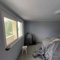 MLB Services - Pokój w trakcie malowania na szaro, zabezpieczone meble folią, widoczny stołek malarski i paczka papierosów na parapecie.