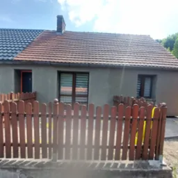 Dom jednorodzinny w trakcie remontu elewacji, widoczne dwie różne dachówki, okna z roletami i drewniane ogrodzenie.