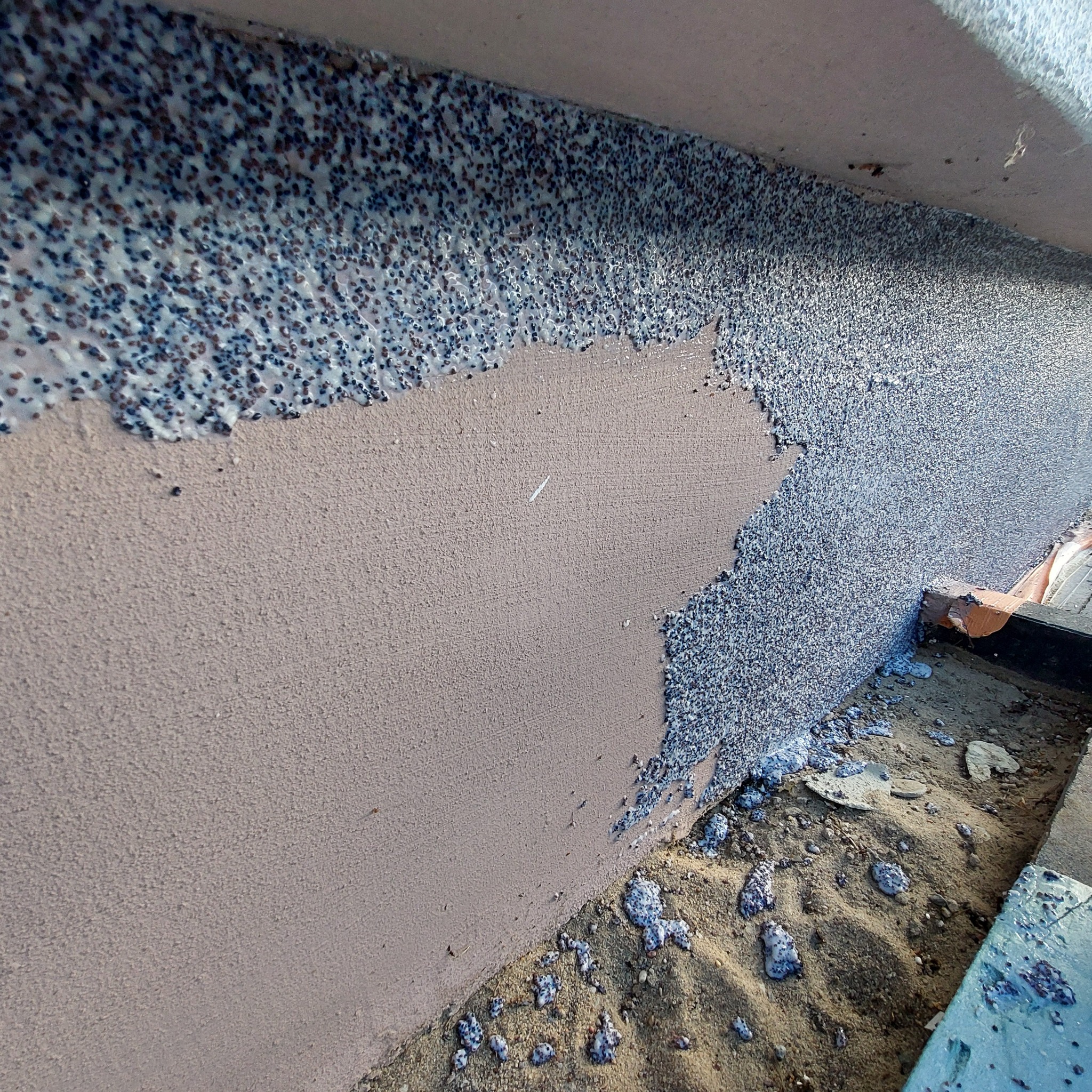 Fragment ściany zewnętrznej z nałożonym tynkiem mozaikowym w trakcie aplikacji, widoczne różnice w fakturze i kolorze pomiędzy nałożonym a surowym tynkiem, na dole resztki tynku na piasku.