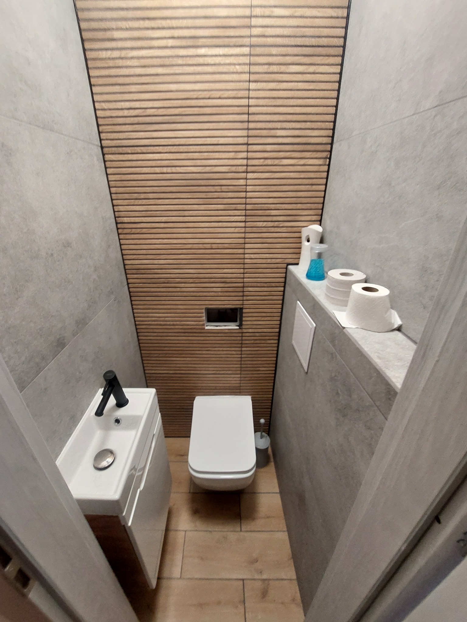 Wąska toaleta z szarymi płytkami na ścianach, drewnianą boazerią, podwieszaną toaletą i małą umywalką z czarną armaturą.