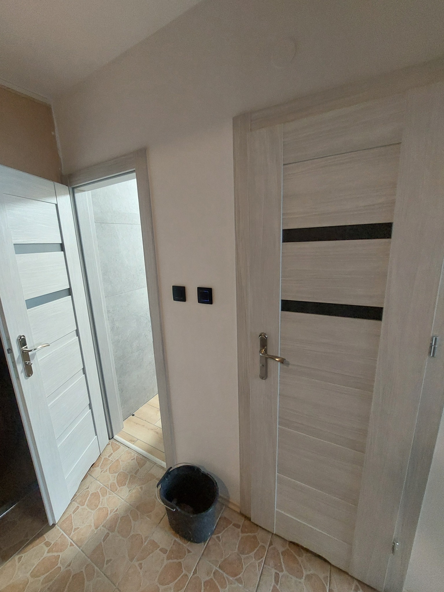 Wnętrze mieszkania w trakcie prac wykończeniowych: widoczne dwie białe, nowoczesne drzwi z poziomymi wstawkami, czarne włączniki światła na ścianie, wiadro na podłodze z terakoty imitującej kamień,...