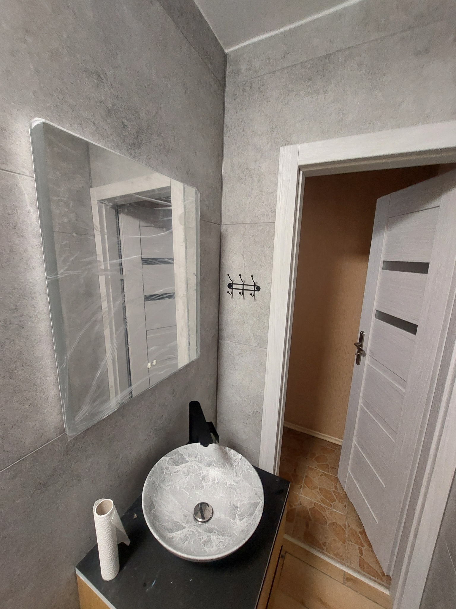 Szara łazienka z umywalką w marmurkowy wzór, czarną baterią, lustrem zabezpieczonym folią, rolką papieru toaletowego i otwartymi drzwiami z widoczną podłogą w innym kolorze.