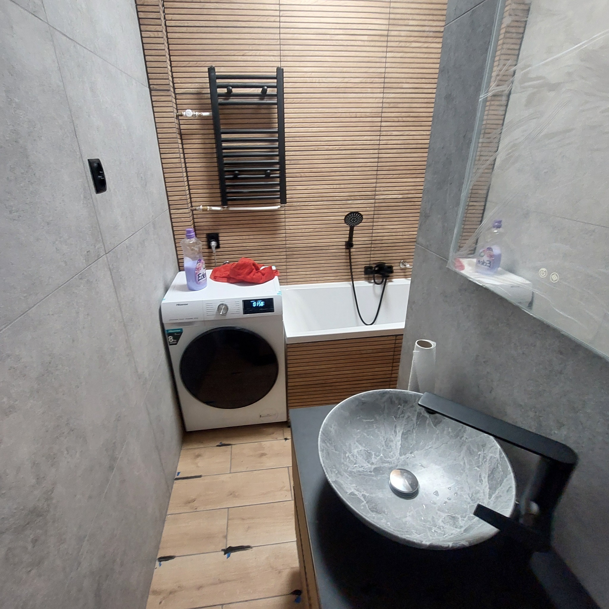 Wykończona łazienka z pralką obok wanny, umywalka marmurkowa z czarną armaturą, drewnopodobne płytki na ścianie i podłodze, grzejnik drabinkowy.