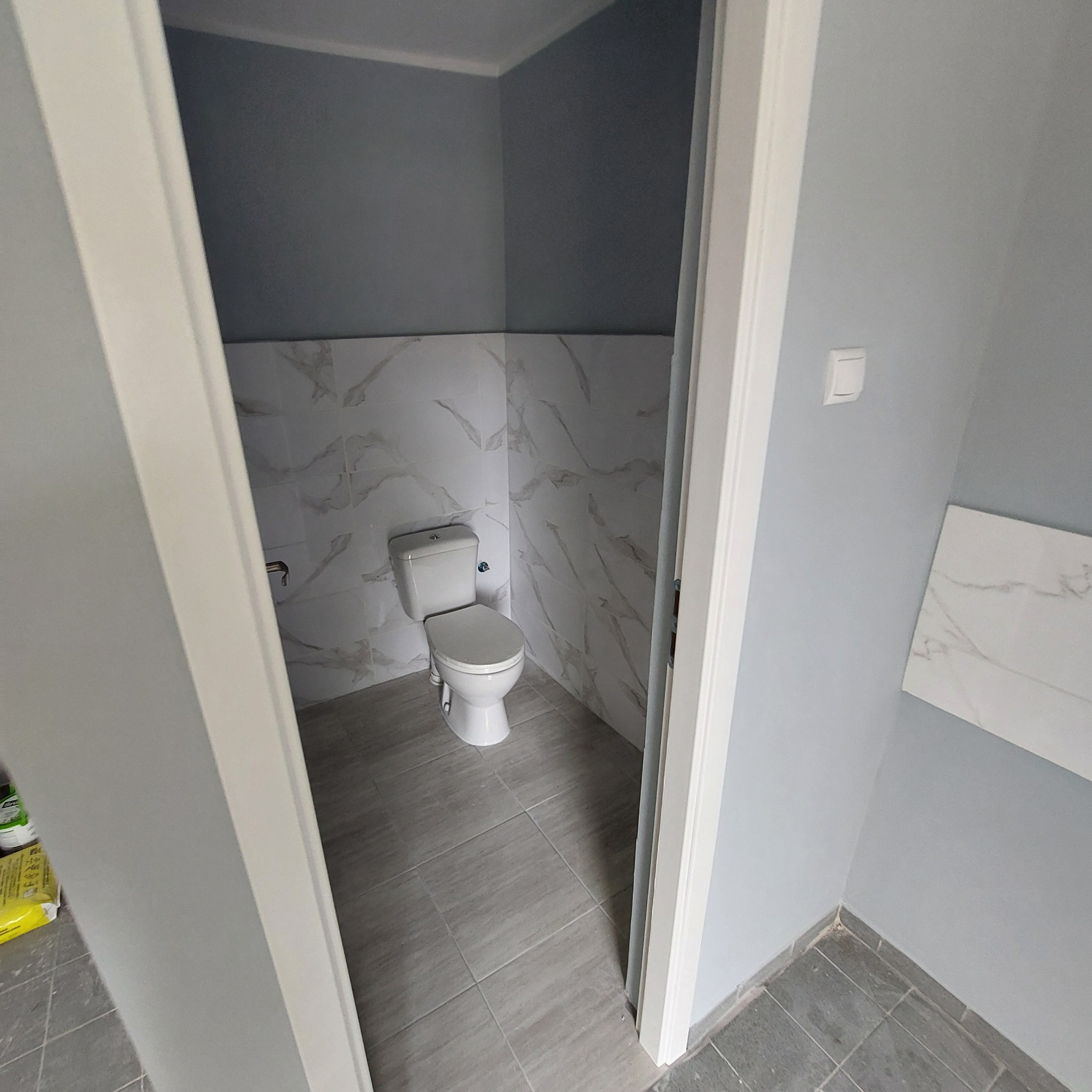 Wnętrze łazienki po remoncie, widok na toaletę, szare płytki podłogowe i ścienne z marmurowym wzorem, pomalowane ściany na szaro, biała armatura, widok z zewnątrz przez białe futryny drzwi.