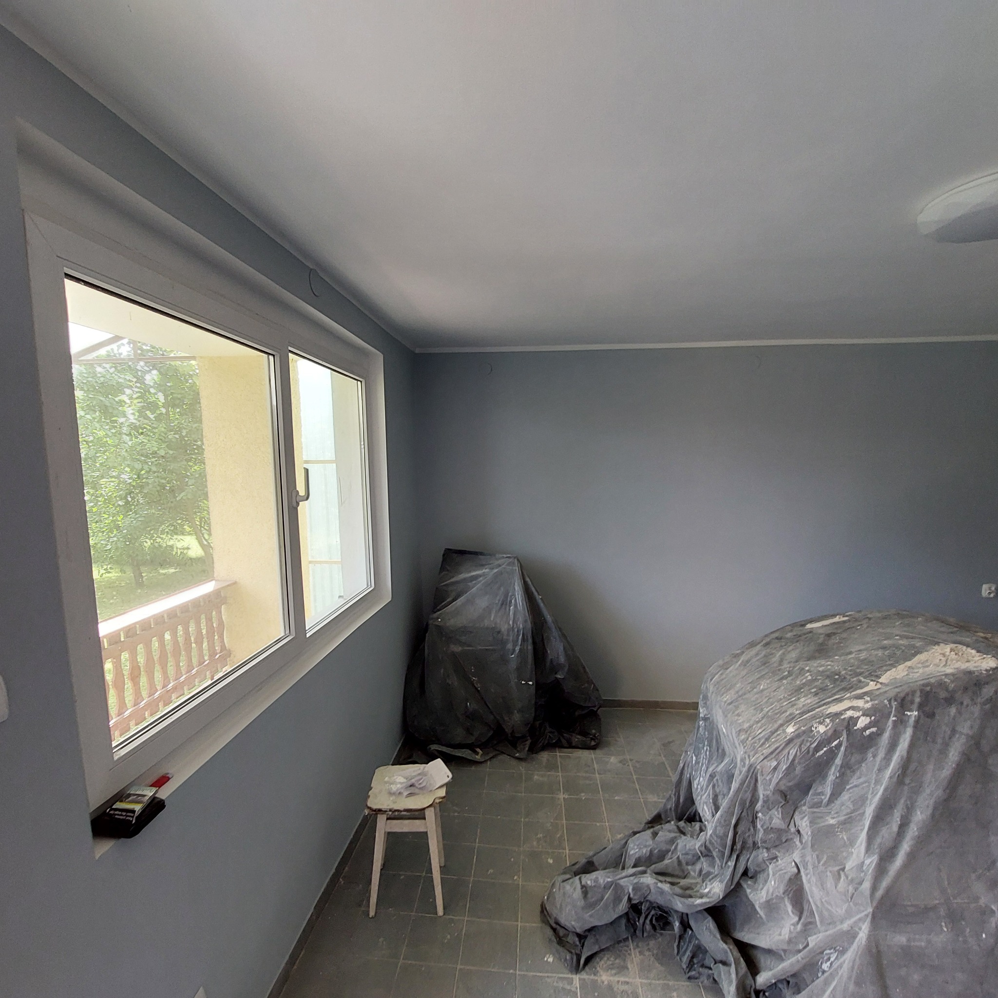 Pokój w trakcie malowania na szaro, zabezpieczone meble folią, widoczny stołek malarski i paczka papierosów na parapecie.