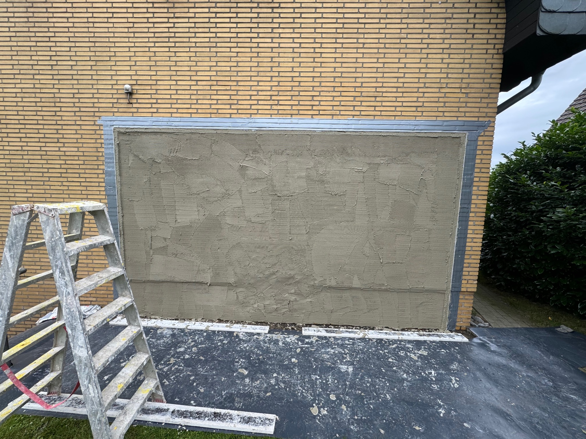 Ściana budynku z żółtej cegły z nałożoną warstwą szarej zaprawy, przygotowana do dalszych prac wykończeniowych, widoczna drabina malarska.