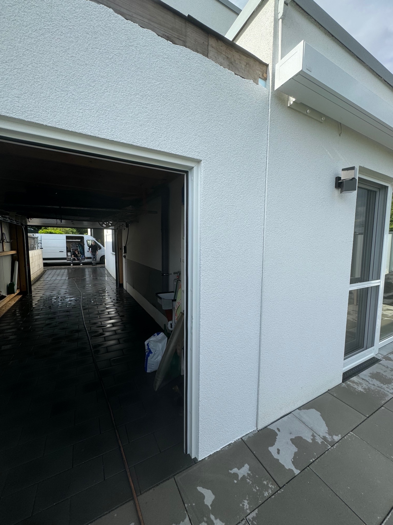 Świeżo otynkowana elewacja garażu z widoczną fakturą, jasny kolor, zadaszenie nad oknem, otwarte wrota garażowe z widocznym wnętrzem i ekipą pracującą w tle.