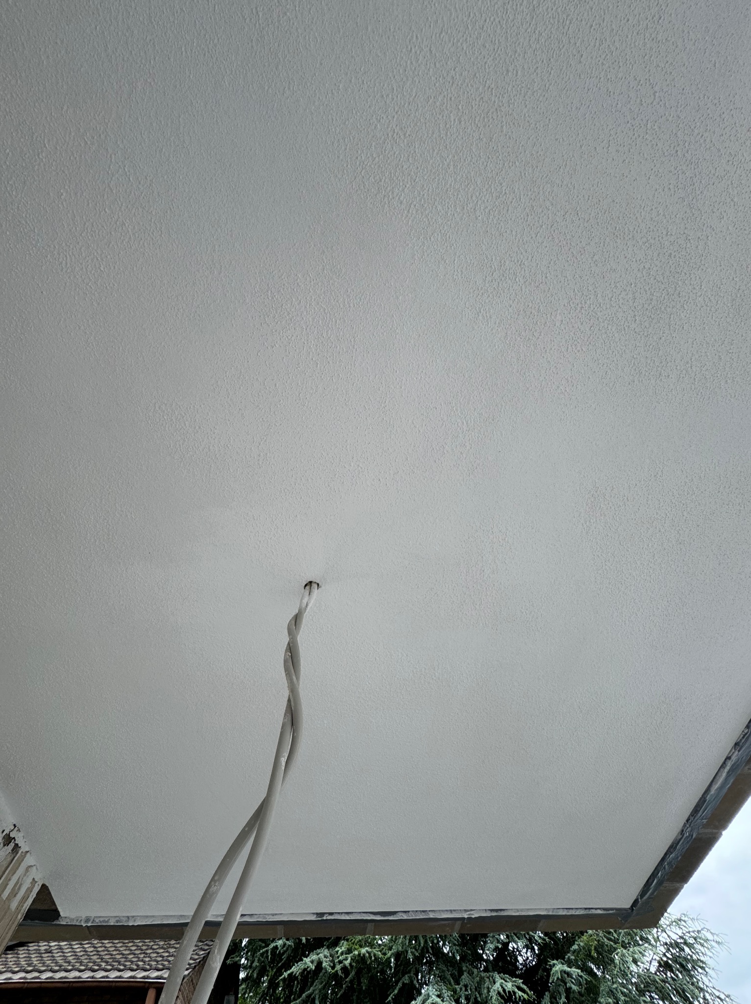 Świeżo nałożona, jasna, strukturalna powłoka na elewacji zewnętrznej budynku, widoczne wystające przewody elektryczne i fragment dachu sąsiedniego budynku.