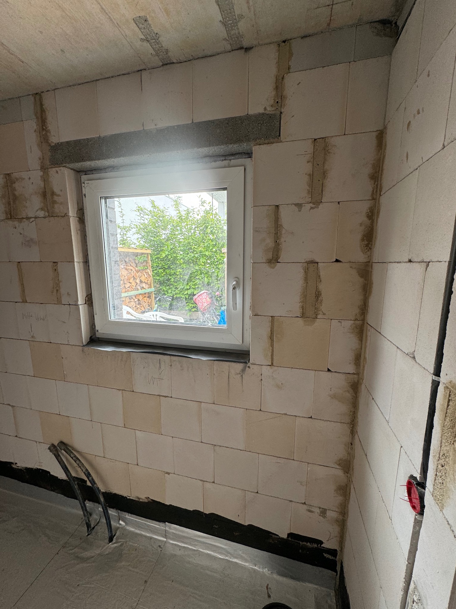 Wnętrze budynku w trakcie budowy: ściany z białych bloczków z widocznymi spoinami, okno z widokiem na zewnątrz, folia na podłodze, instalacja elektryczna w ścianie.