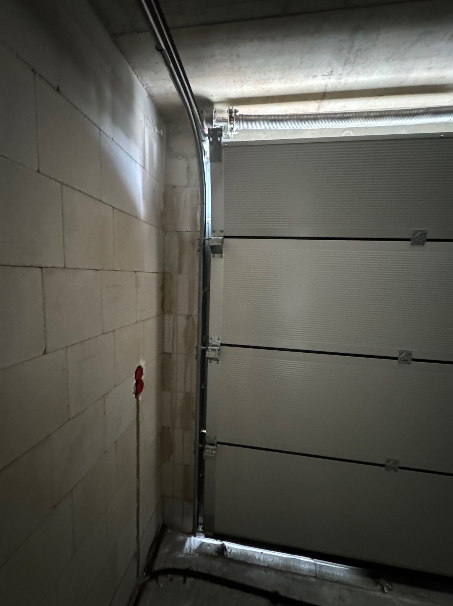 Wnętrze garażu z widoczną ścianą z betonu komórkowego, szyną prowadzącą i segmentową bramą garażową w trakcie montażu, z widocznymi elementami mocującymi i sprężyną naciągową.