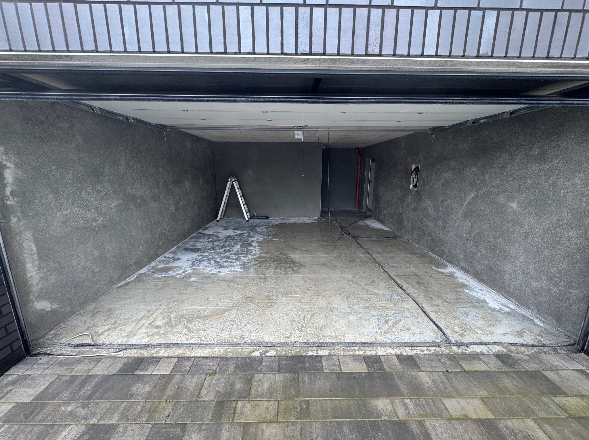 Wnętrze garażu po nałożeniu szarego tynku, widoczna drabina oparta o ścianę, ślady po pracach na posadzce i instalacje pod sufitem.