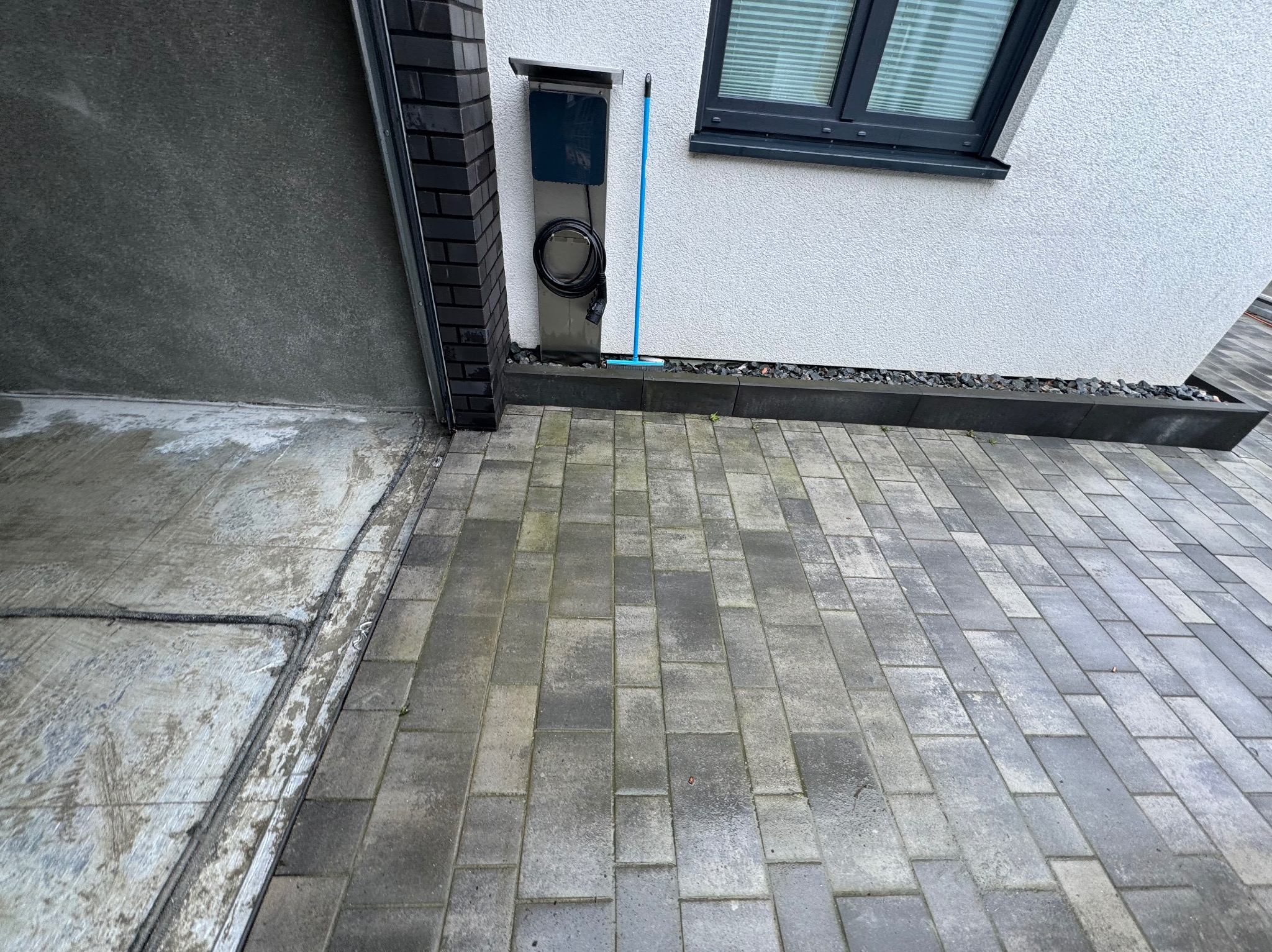 Fragment elewacji domu z szarą kostką brukową na podjeździe, widoczna ściana o szorstkiej fakturze, okno z szarymi ramami, czarna cegła klinkierowa i skrzynka z kablem do ładowania pojazdu...